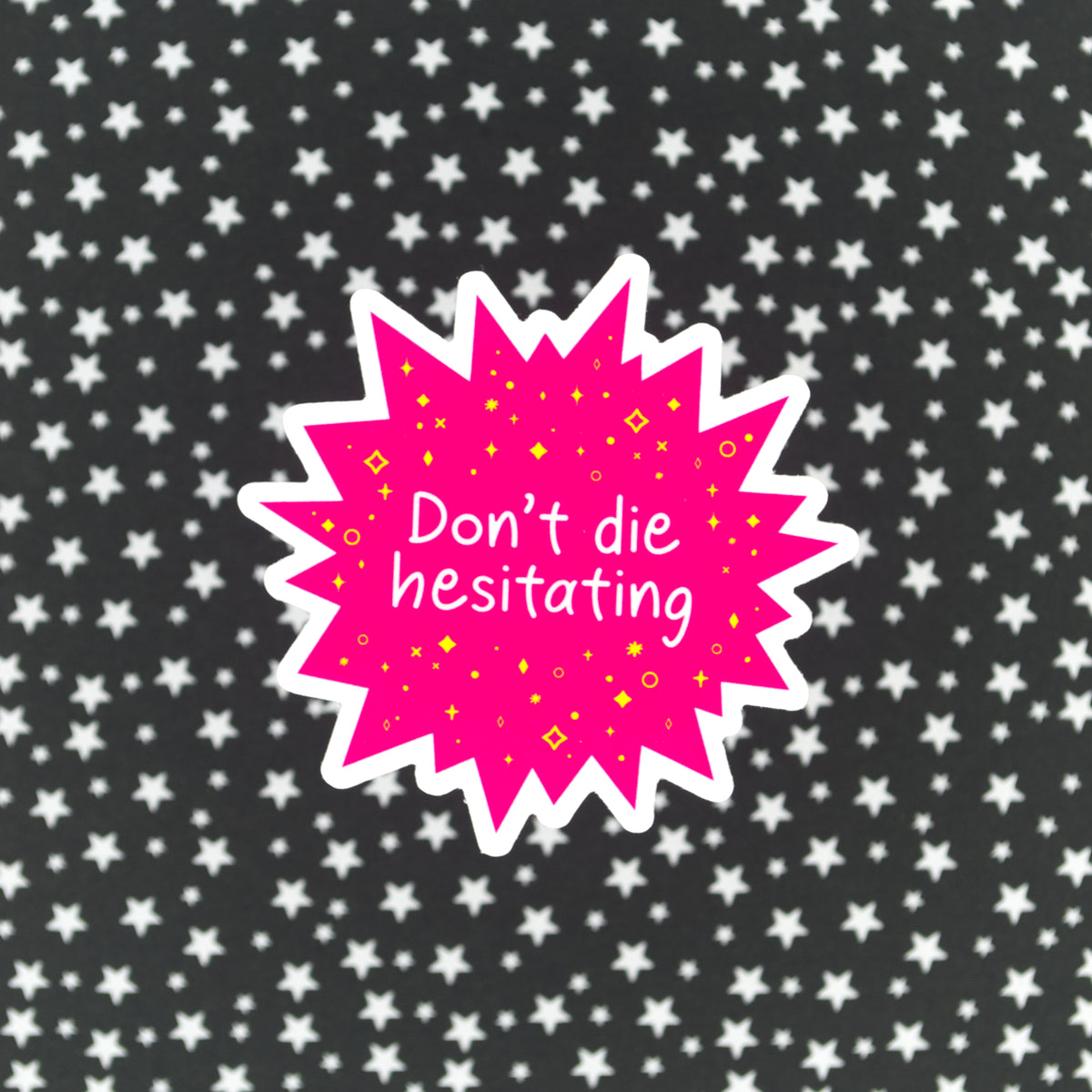 A bright pink star-burst shaped sticker with the words "Don't die hesitating" in white handwriting surrounded by yellow stars and sparkles. The sticker sits against a black background with silver stars