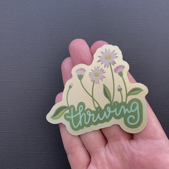 A video of a person's hand rotating the Thriving sticker so you can see the metallic effect.