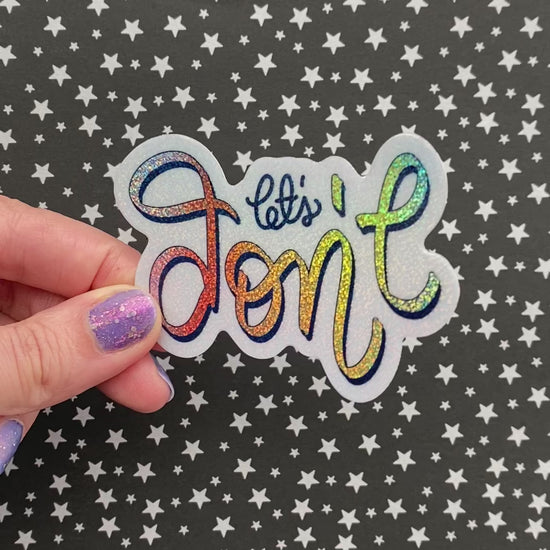 A person with purple nail polish holding a rainbow glitter sticker to show how the light hits the sticker. The sticker is held againt a black background with silver stars.