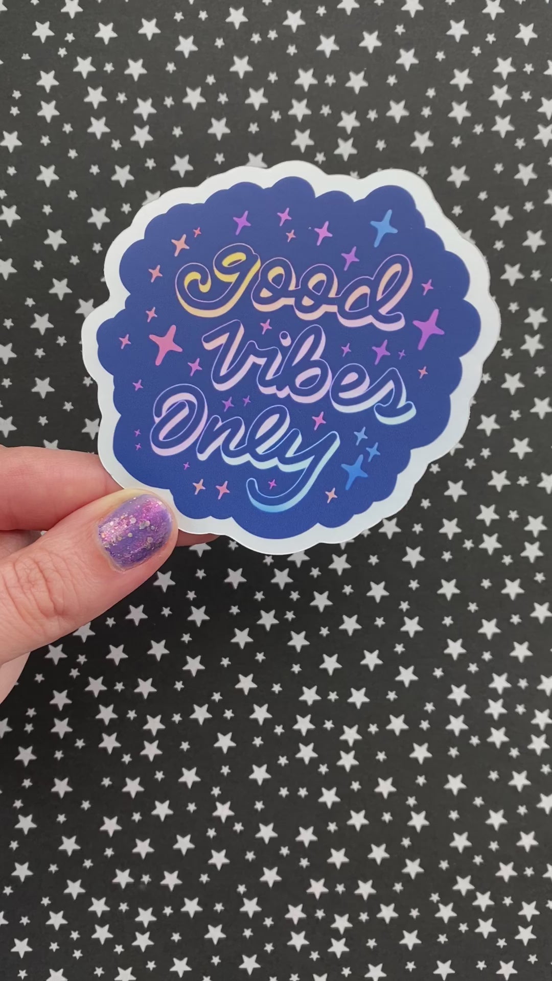 A video of a person holding a blue holographic sticker with the words "good vibes only" written in calligraphy and surrounded by rainbow sparkles. The person rotates the sticker to show its holographic finish.
