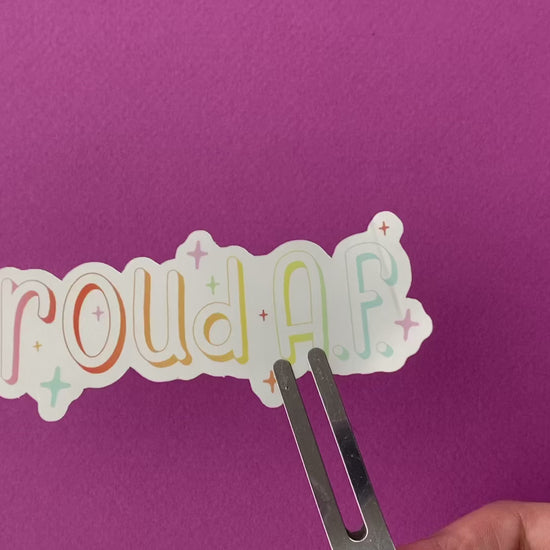 A clip holds a metallic silver sticker against a bright purple paper background. The sticker says "proud A.F." in a pastel rainbow gradient and is surrounded by sparkles. As the sticker rotates, the metallic surface reflects lightly at different angles.
