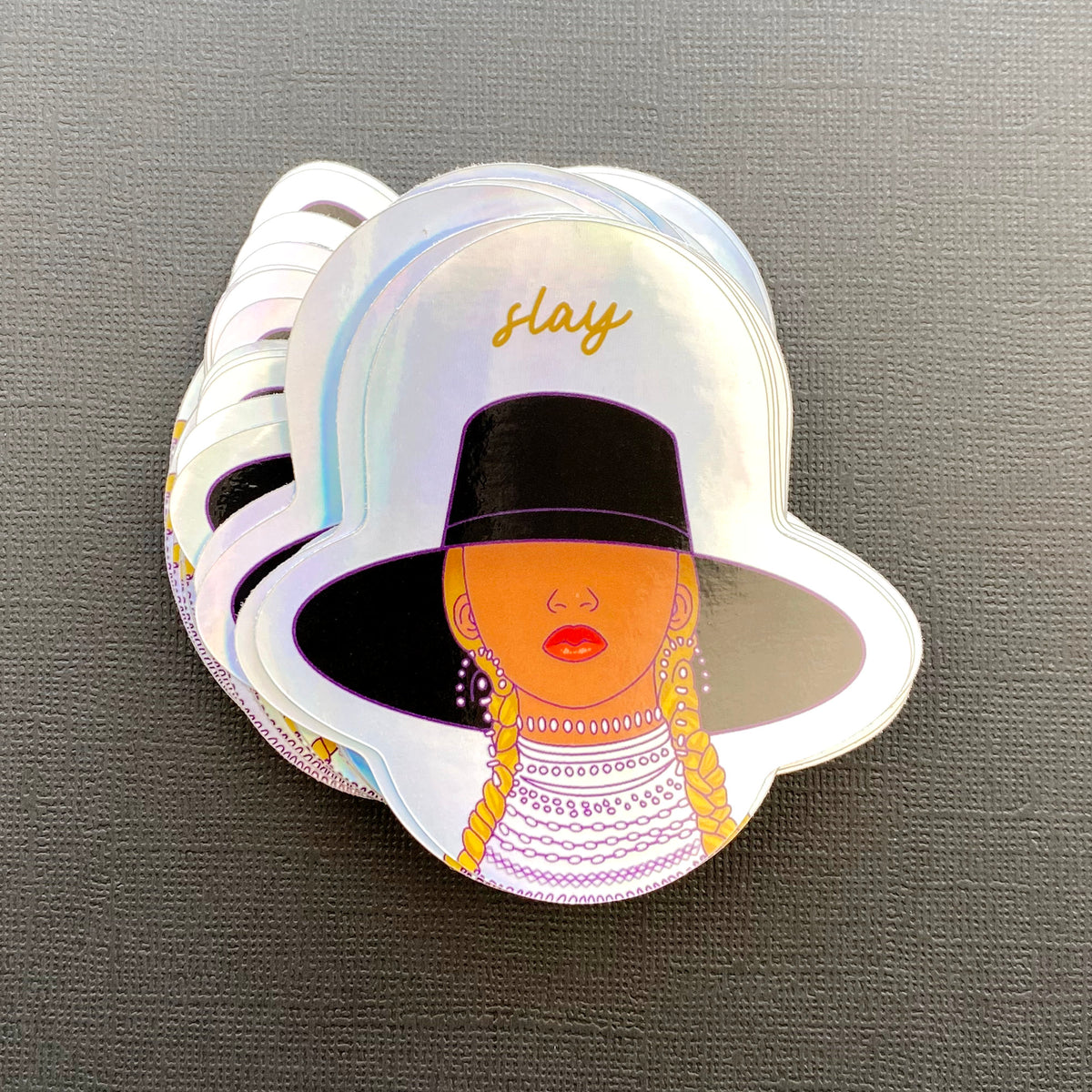 A photo of a holographic sticker illustrated with a portrait of Beyonce from the Formation video and the word "slay" over her head. The sticker sits on a black paper background.