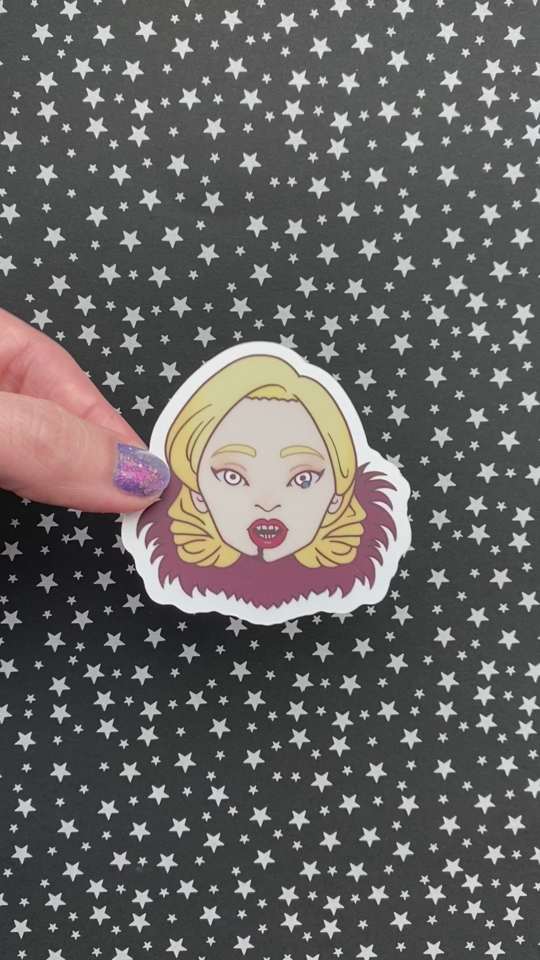 A person with purple nail polish holding an illustrated sticker of Lady Gaga from American Horror Story to show it's holographic material. The background is black paper with silver stars.