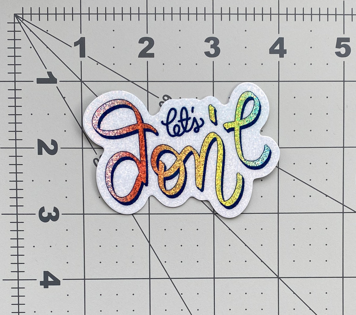 A rainbow glitter sticker that says "let's don't" in calligraphy and sits on a ruled background to show it's size.